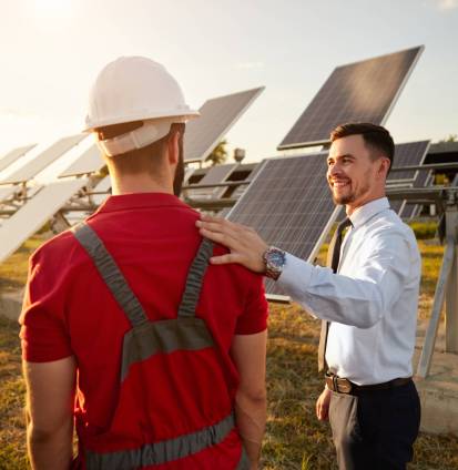 Male investor patting technician in uniform on shoulder while standing near photovoltaic panels in countryside and looking at each other