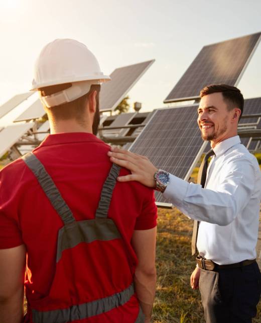 Male investor patting technician in uniform on shoulder while standing near photovoltaic panels in countryside and looking at each other